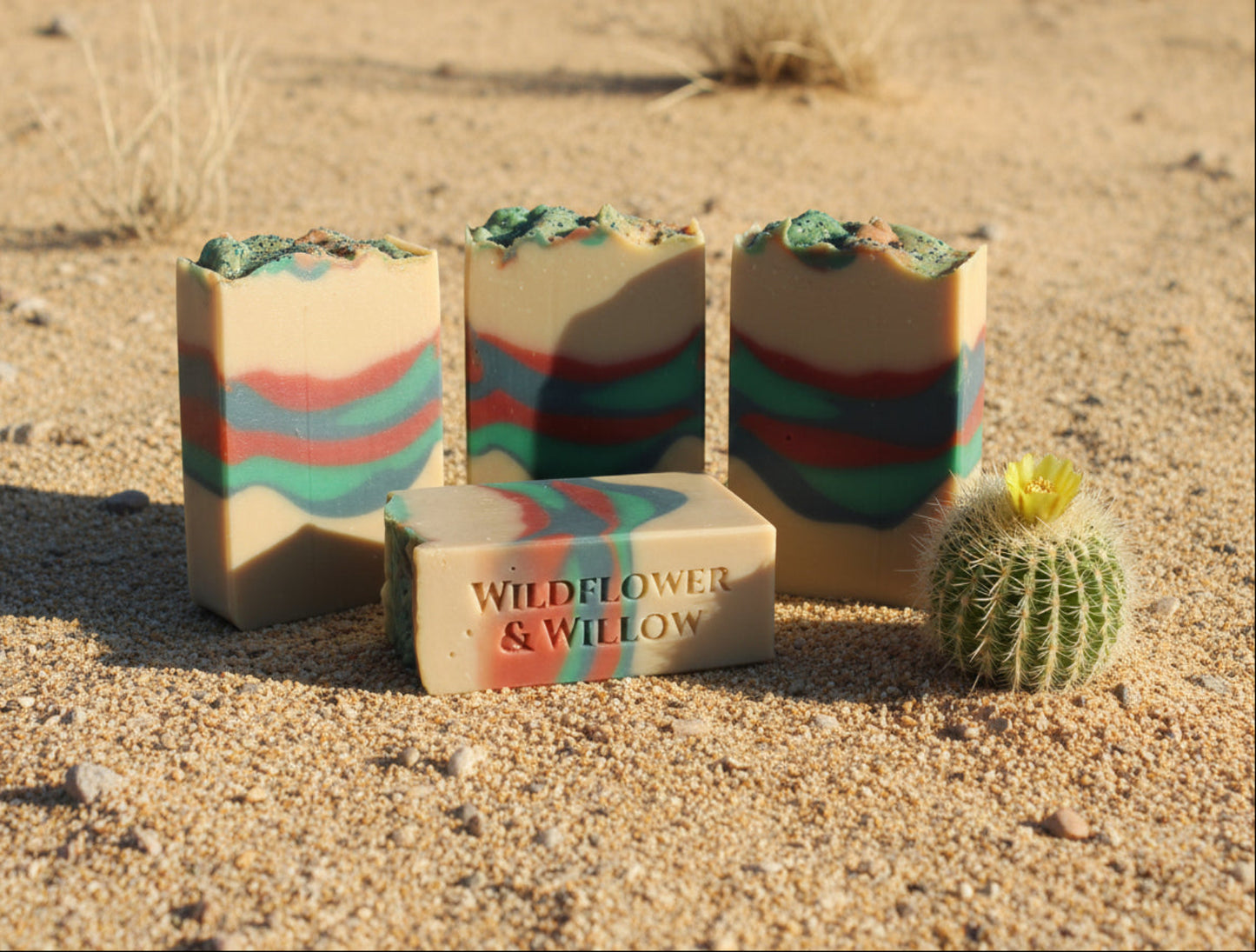 4 bars of multicolored soap in desert sand with a small cactus