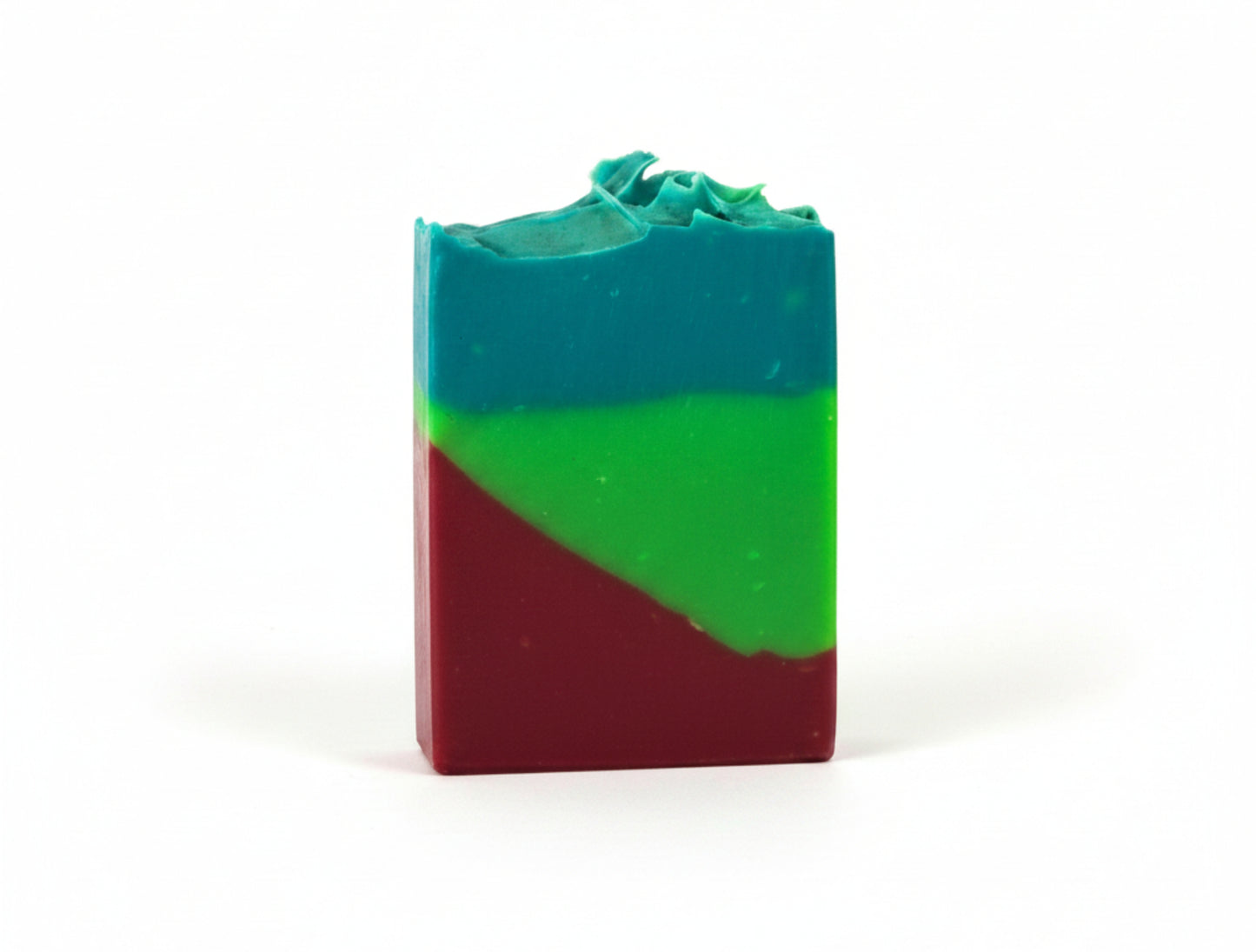 A handmade rectangular bar of soap with a three-color layered design of teal blue, green, and deep red, standing upright on a white surface against a white wall.
