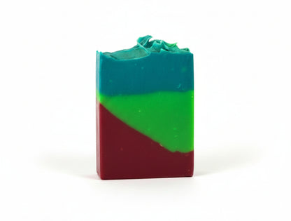 A handmade rectangular bar of soap with a three-color layered design of teal blue, green, and deep red, standing upright on a white surface against a white wall.